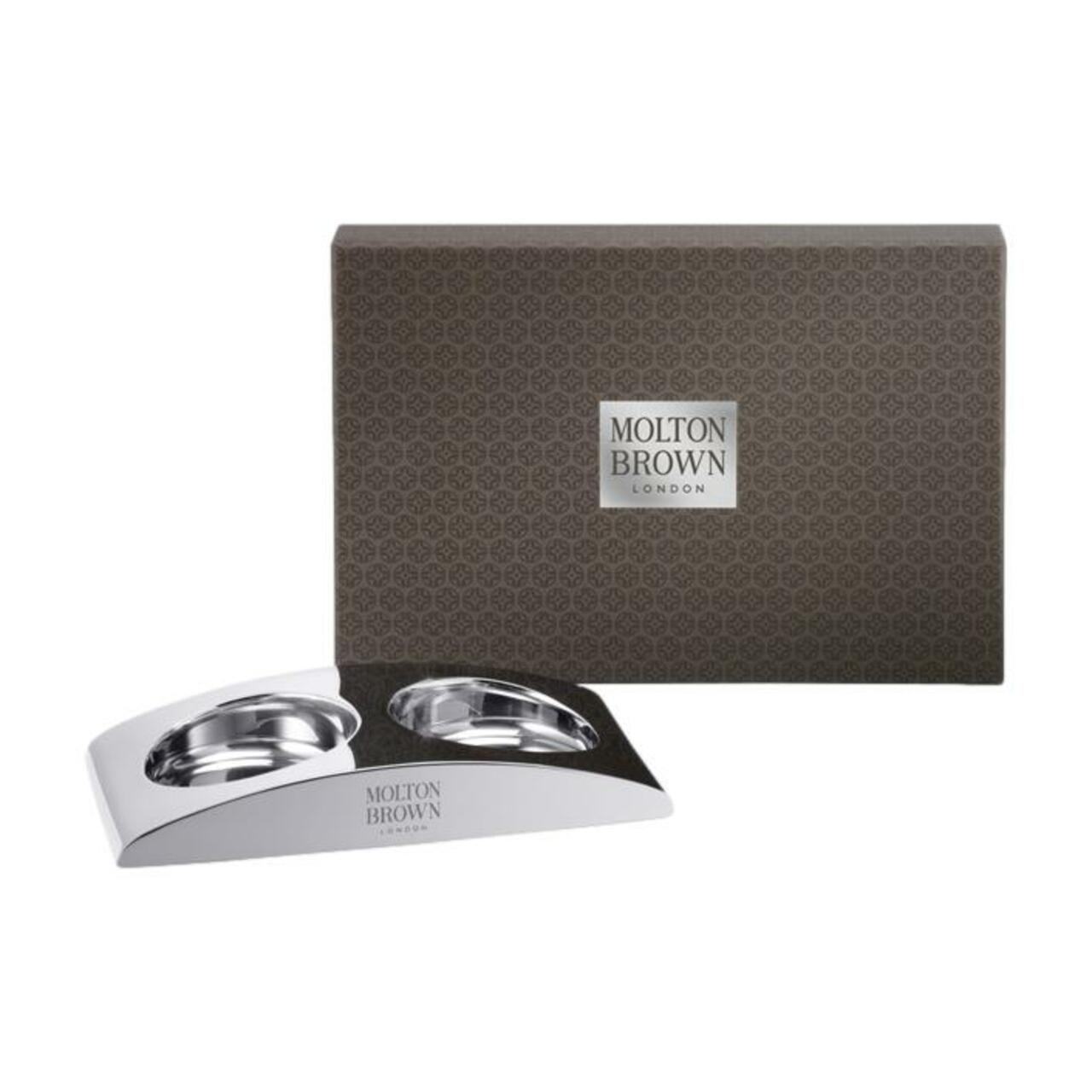 Silberfarbene Halterung mit zwei Vertiefungen, daneben eine braune Schachtel mit Molton Brown Logo. Verpackung.