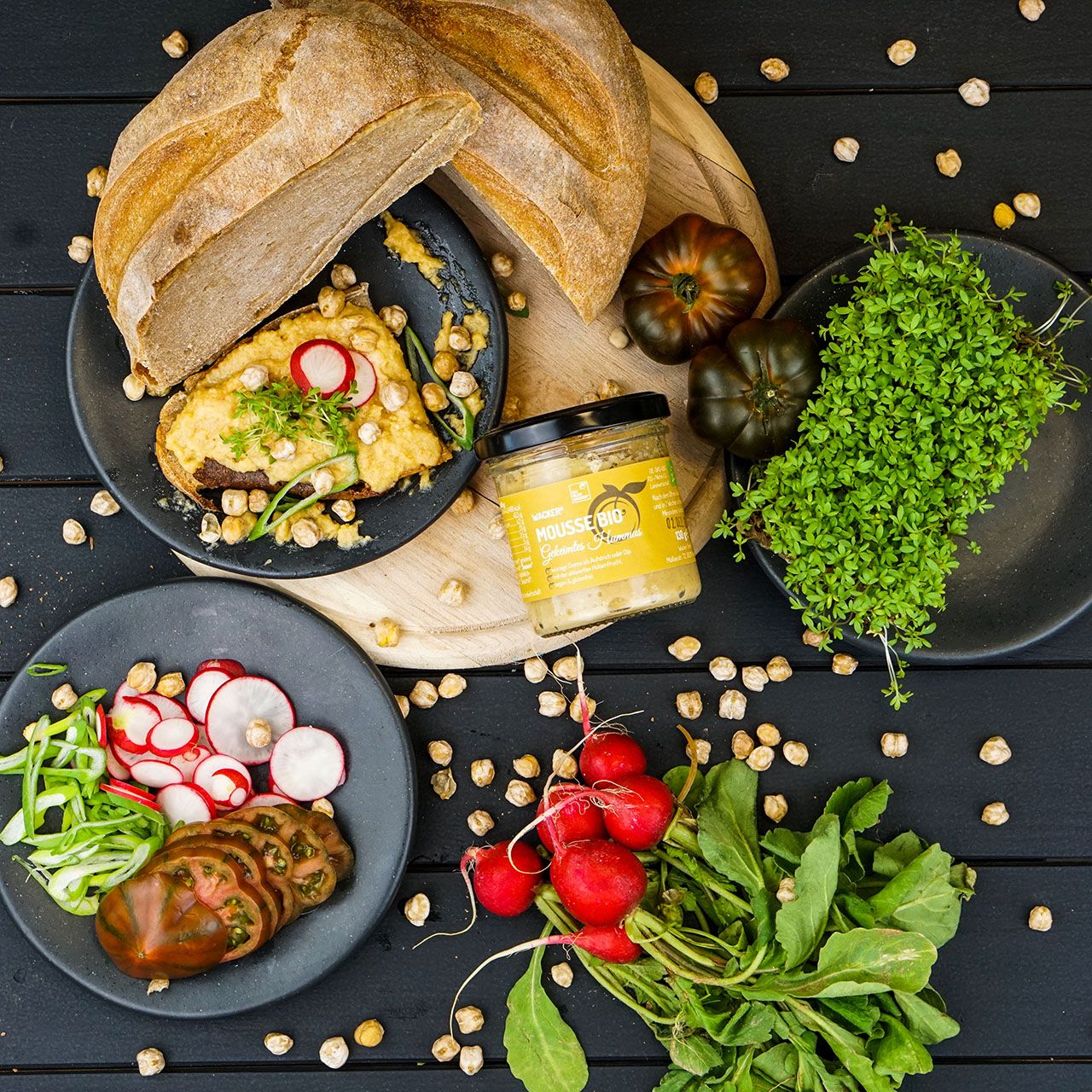 Hummus-Glas neben Brot, Gemüse und Kichererbsen. Aufschrift: Mousse Bio.