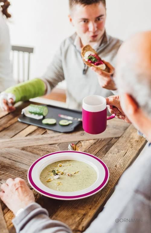 Personen am Tisch. Ein Teller mit Suppe, ein Becher und ein Brot. ORNAMIN-Logo.