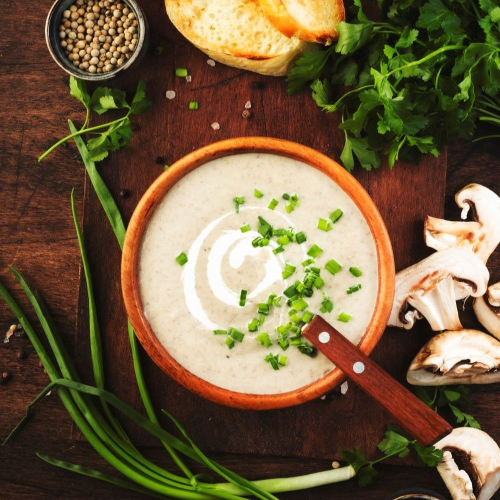 Holzschale mit cremiger Suppe, garniert mit Schnittlauch und Sahne. Daneben Champignons, Brot, Kräuter und Pfefferkörner.