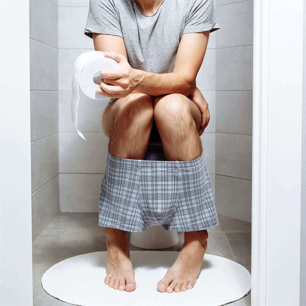 Person auf Toilette, hält Toilettenpapier. Unterhose mit Karomuster. Füße auf weißer Matte.