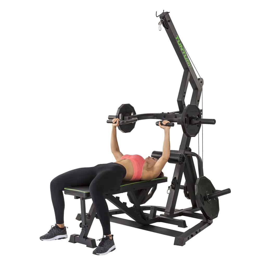 Fitnessgerät mit Hantelstange und Gewichten. Eine Person liegt auf einer Bank und hebt die Hantelstange. Schwarze Konstruktion mit grünen Akzenten.
