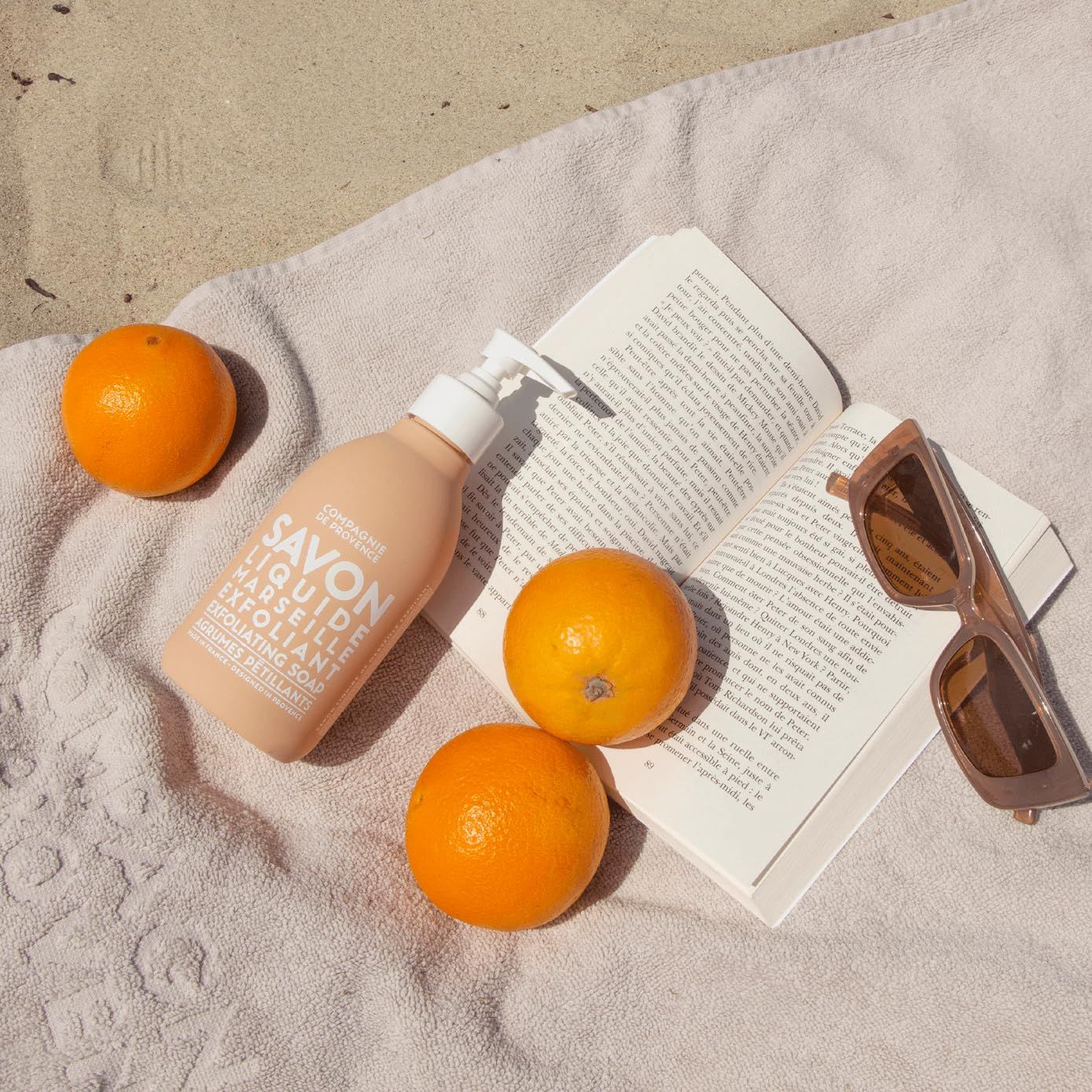 Flüssigseife, Orangen, Buch und Sonnenbrille auf einem Handtuch am Strand.