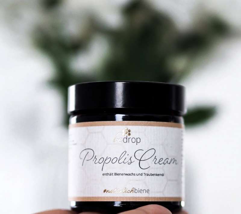 Glasdose mit "Propolis Cream" und schwarzem Deckel. "be drop" Logo. Enthält Bienenwachs und Traubenkernöl. Hand hält Dose.
