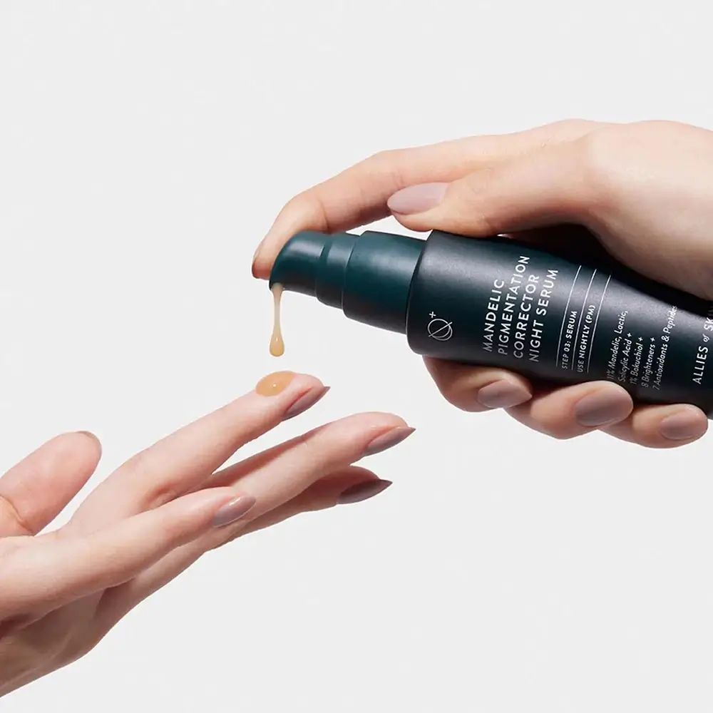Hände halten Serumflasche. Serum tropft auf die Hand. Aufschrift: Mandelic Pigmentation Corrector Night Serum.