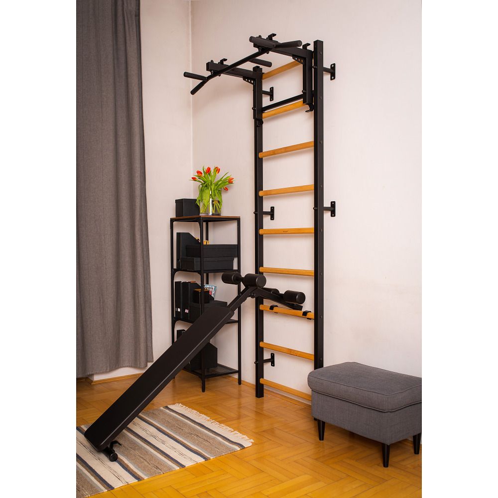 Fitness-Sprossenwand mit Zubehör. Eine schwarze Bank steht vor der Wand. Blumen und ein Regal sind zu sehen.