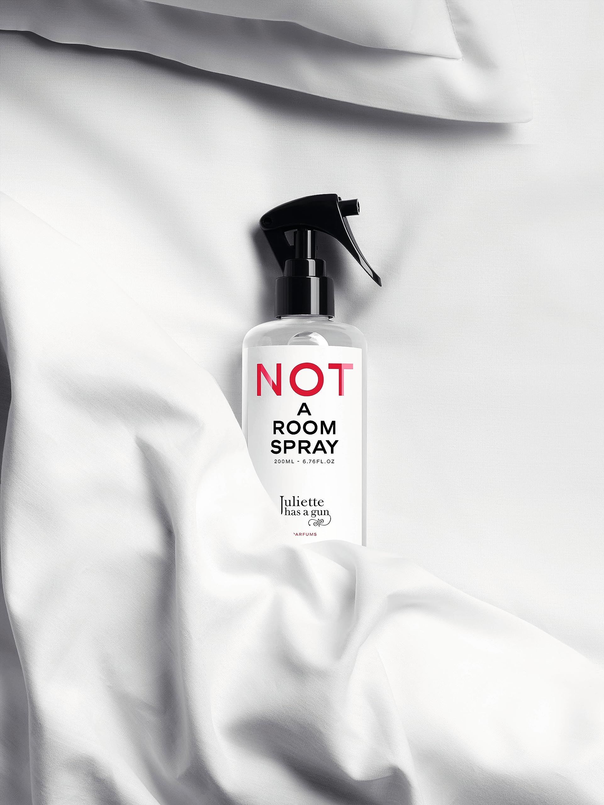 Transparente Flasche mit schwarzem Sprühkopf auf weißem Stoff. Roter Schriftzug: NOT A ROOM SPRAY. Marke: Juliette has a gun.