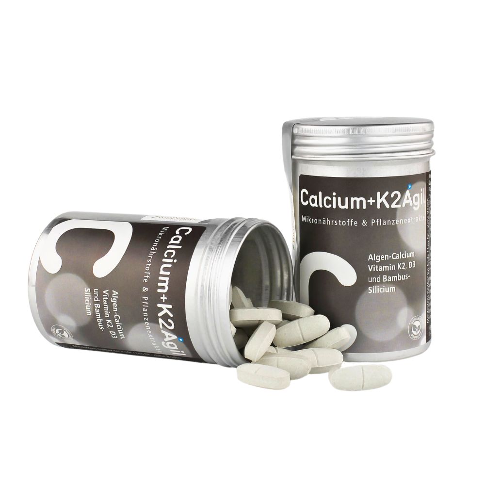 Zwei silberne Dosen mit Tabletten. Auf den Dosen steht "Calcium+K2Agil". Tabletten liegen verstreut.