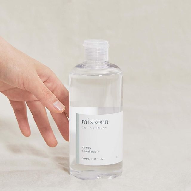 Hand neben Flasche mit weißem Etikett. Aufschrift: mixsoon Centella Cleansing Water. Produkt auf hellem Hintergrund.