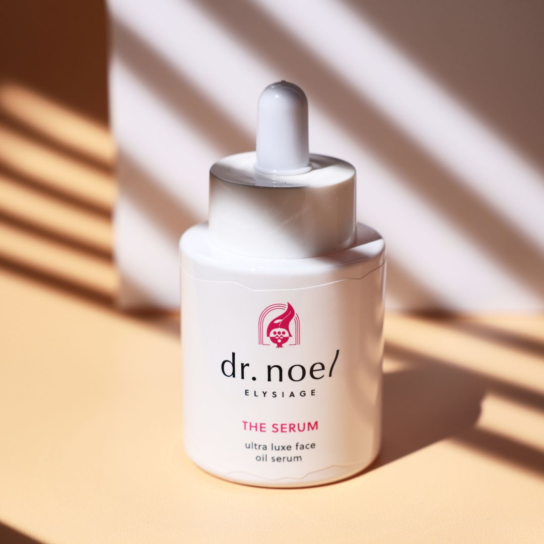 Weiße Serumflasche. Aufschrift: dr. noel/ ELYSIAGE, THE SERUM, ultra luxe face oil serum. Rosa Akzente. Hintergrund: hellbraun.