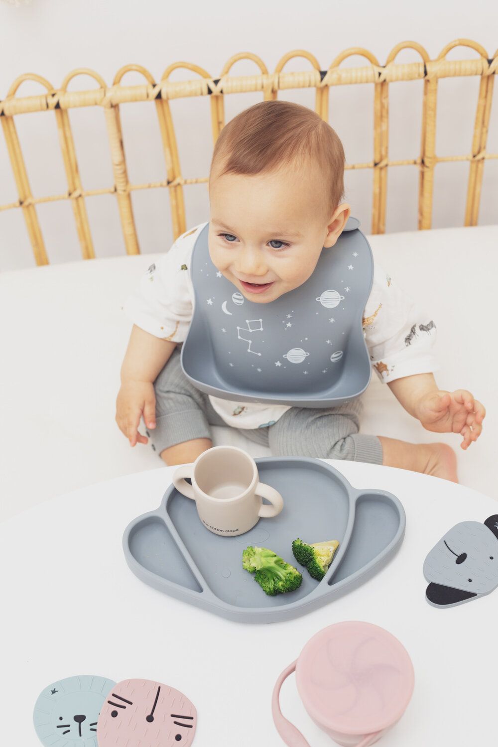 Baby sitzt am Tisch mit Teller, Becher und Lätzchen. Teller in Bärenform mit Brokkoli. Lätzchen mit Weltraum-Motiv.