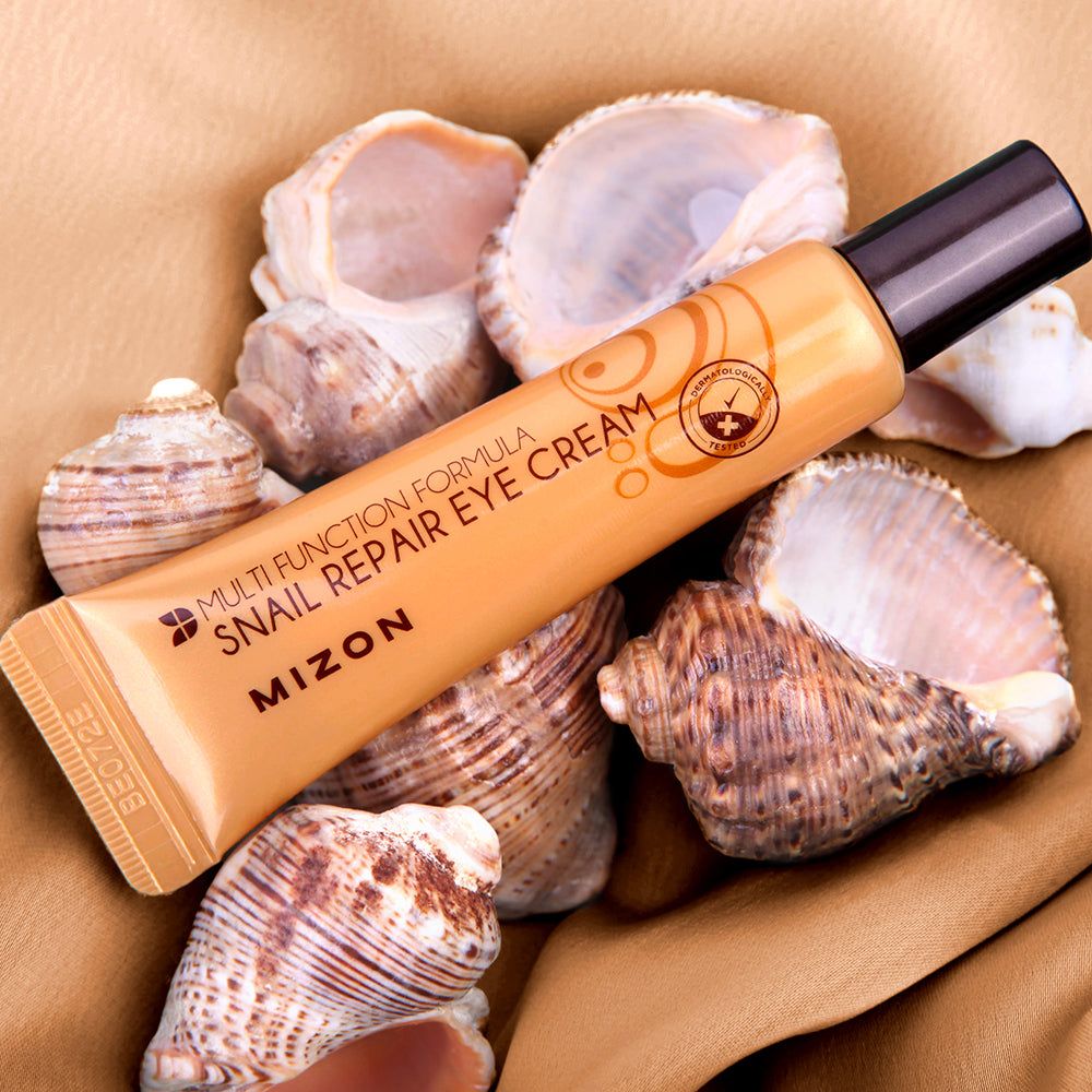 Goldfarbene Tube mit schwarzem Verschluss, auf Muscheln. Aufschrift: Mizon Snail Repair Eye Cream.