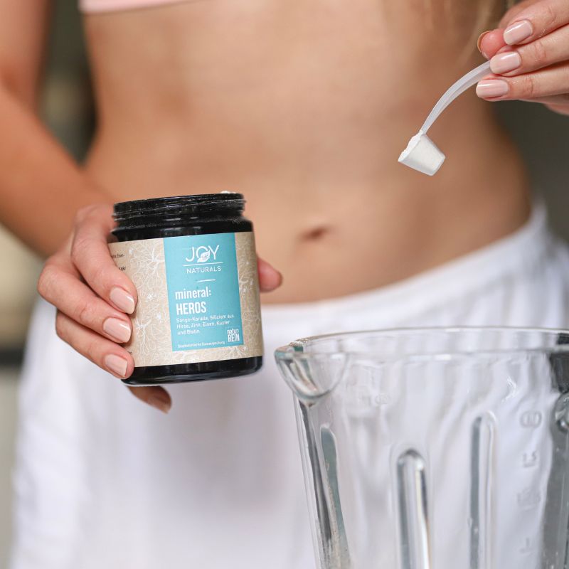 Person hält eine Dose JOY NATURALS und einen Messlöffel. Im Hintergrund ein Mixer.