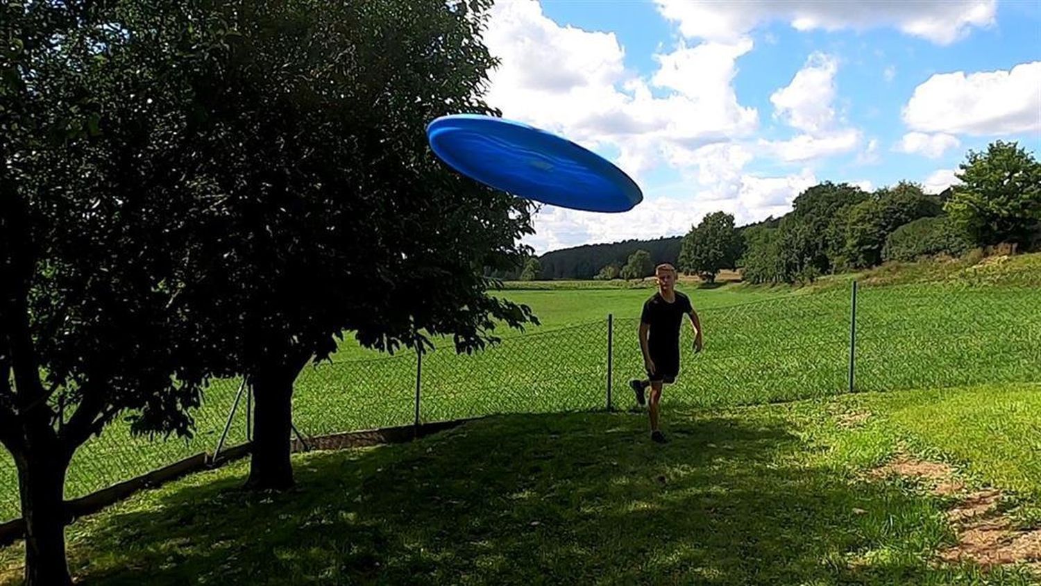 Blaue Wurfscheibe im Flug. Person rennt im Hintergrund.