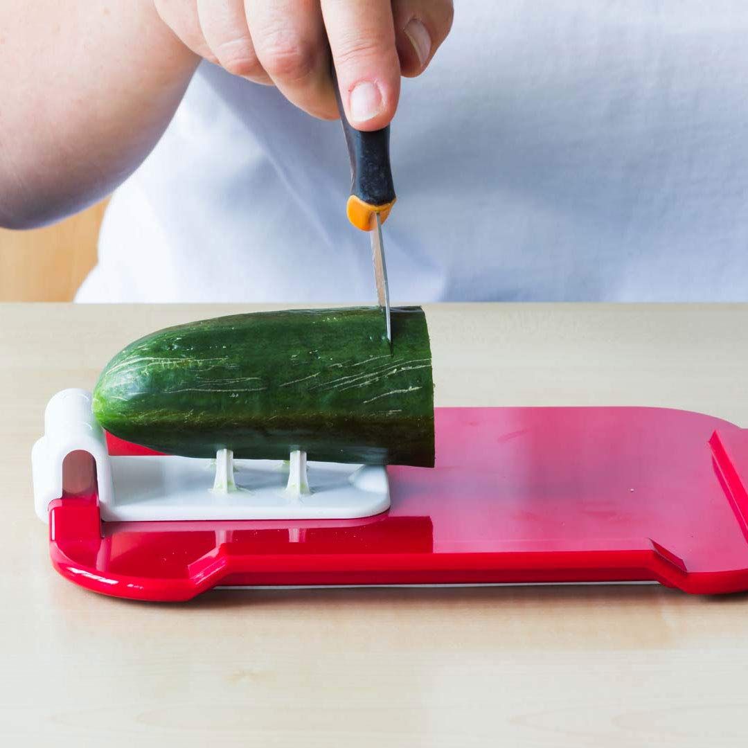 Rotes Essbrettchen mit Gurke und Messer. Gurke wird geschnitten. Weiße Halterung mit Stiften.