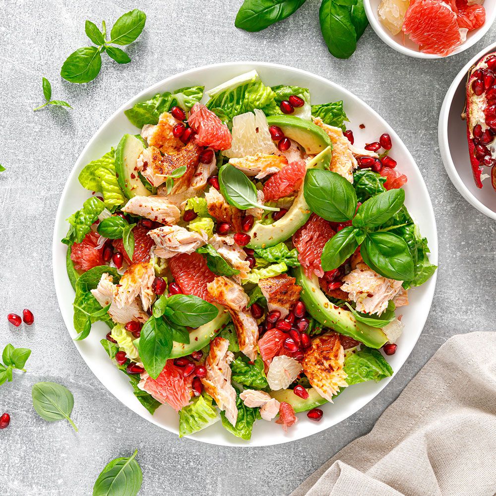 Salat mit Lachs, Avocado, Grapefruit, Granatapfel und Basilikum. Auf weißem Teller, mit grauem Hintergrund.