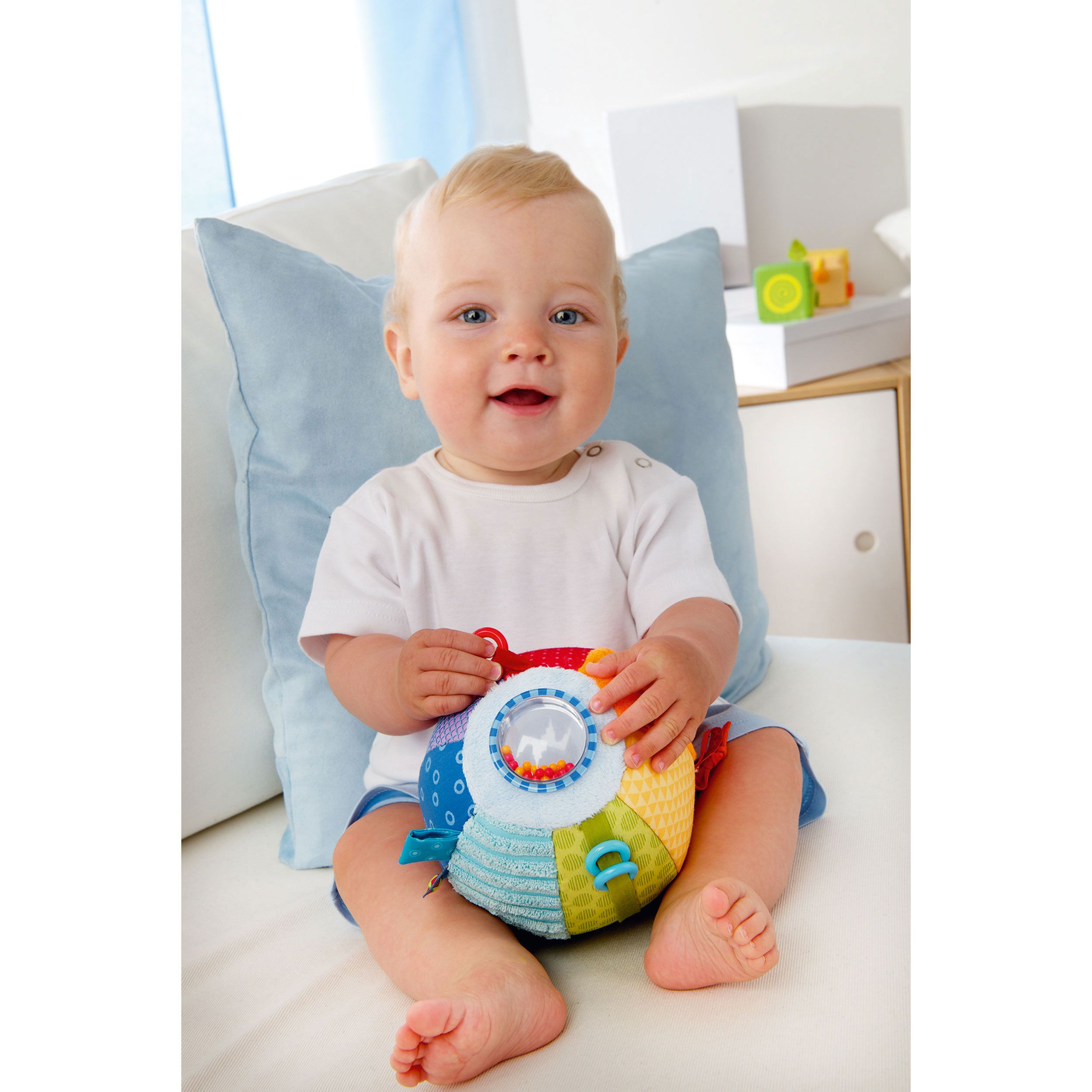 Baby hält bunten Greifball. Der Ball hat verschiedene Texturen und Farben. Baby sitzt auf einer Couch.