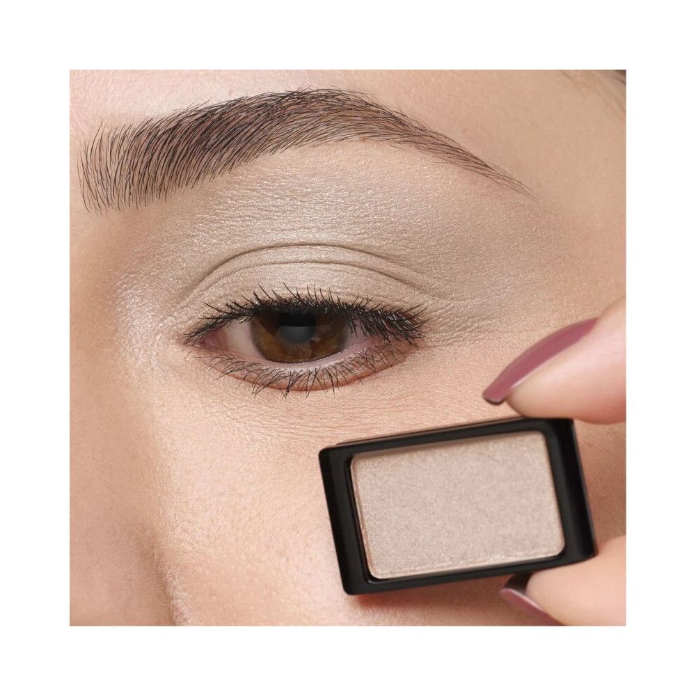 Ein Augenlid-Schatten in einem schwarzen Behälter. Die Hand hält das Produkt neben einem Auge, auf dem der Lidschatten aufgetragen wurde.