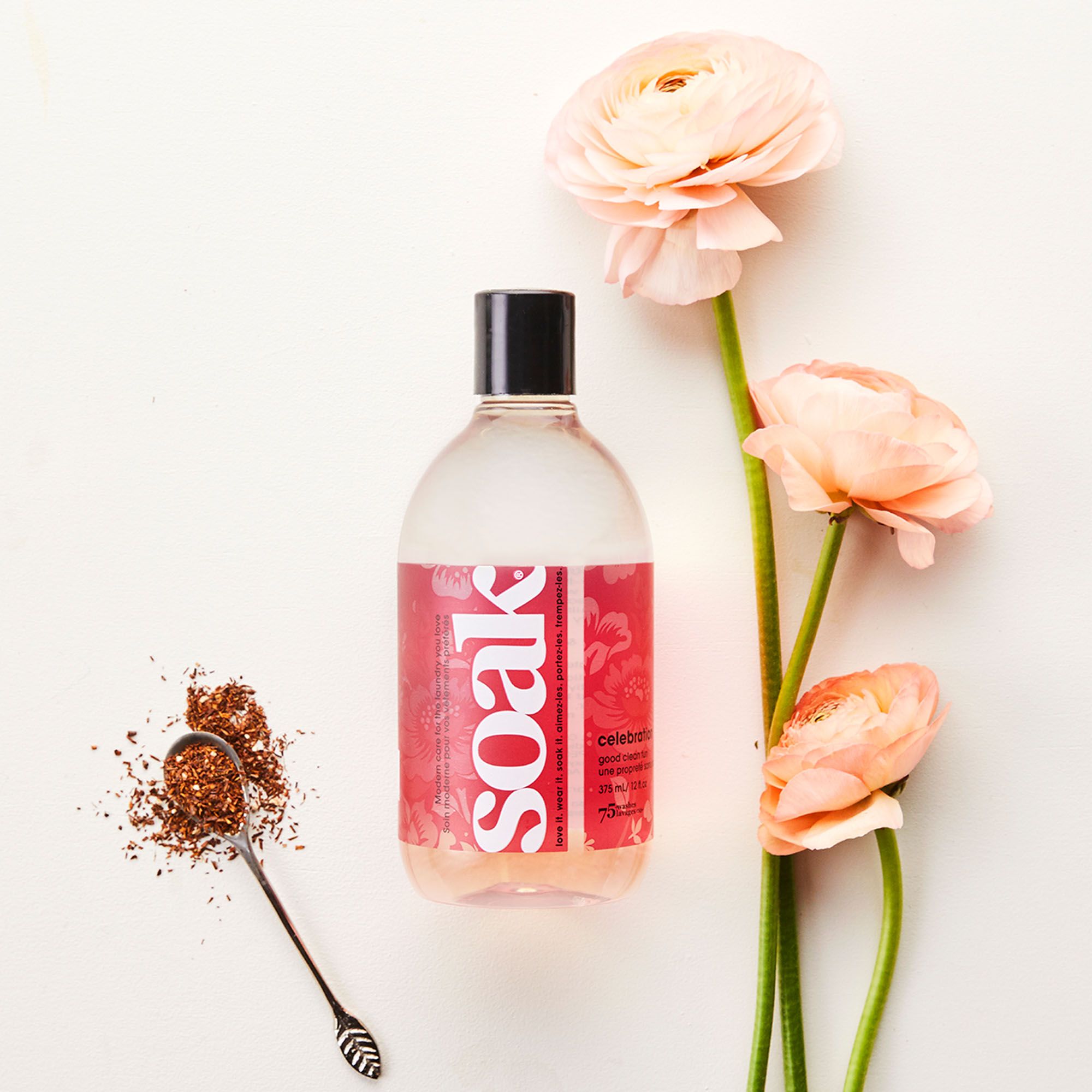 Klare Flüssigkeit in einer Flasche mit roter Etikette. "Soak"-Logo. Daneben Blüten und Teeblätter auf weißem Hintergrund.