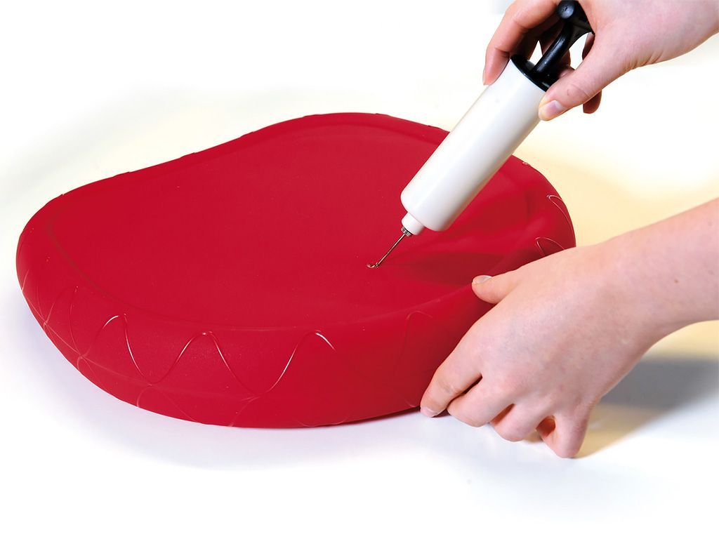 Eine Hand pumpt Luft in ein rotes Sitzkissen mit einer Pumpe. Das Kissen hat eine runde Oberseite und eine wellenförmige Unterseite.