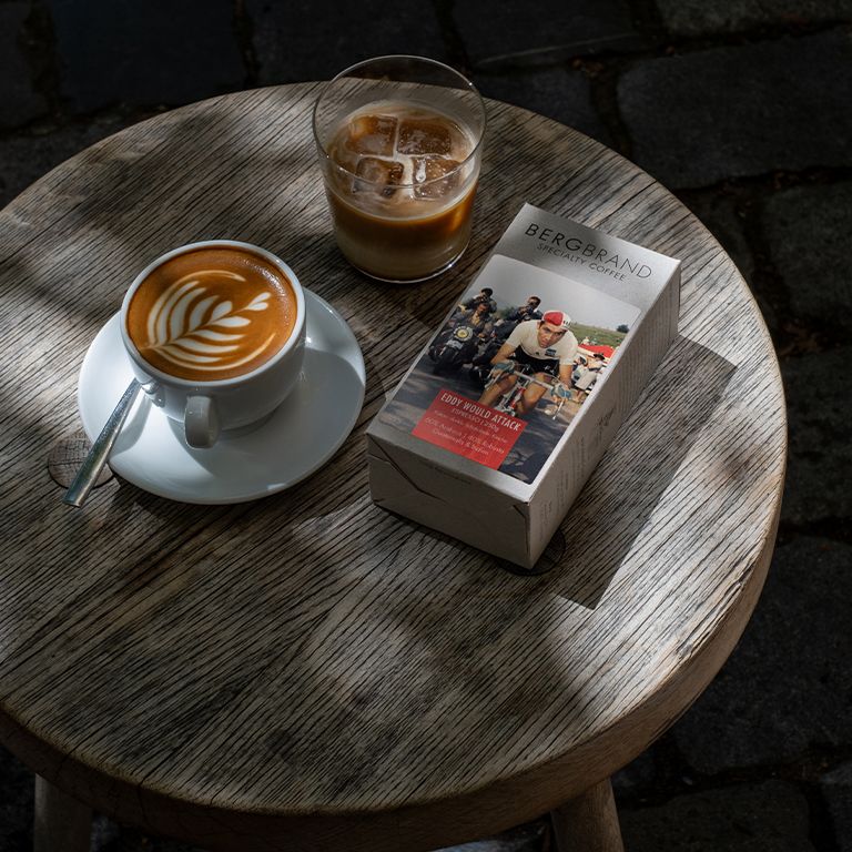 Kaffeetasse mit Latte Art, Kaffeepackung und Getränk mit Eiswürfeln auf einem Holztisch. BERGBRAND Eddy would Attack.
