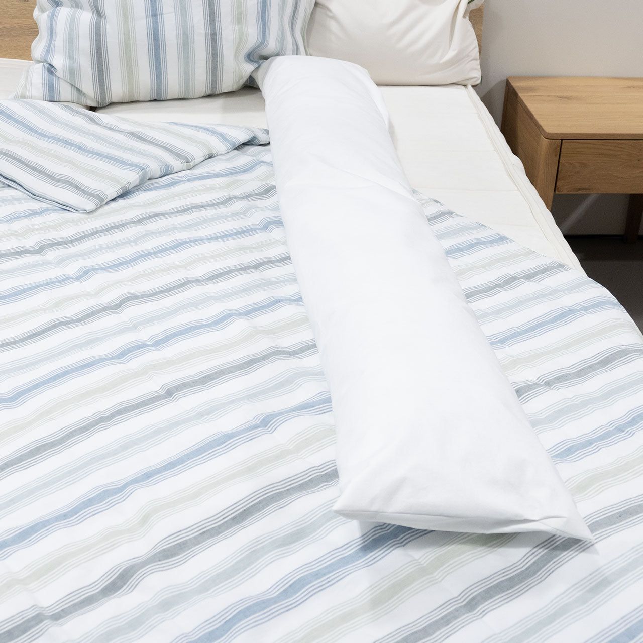 Weißes Seitenschläferkissen auf einem Bett. Kissen liegt längs auf gestreifter Bettwäsche.