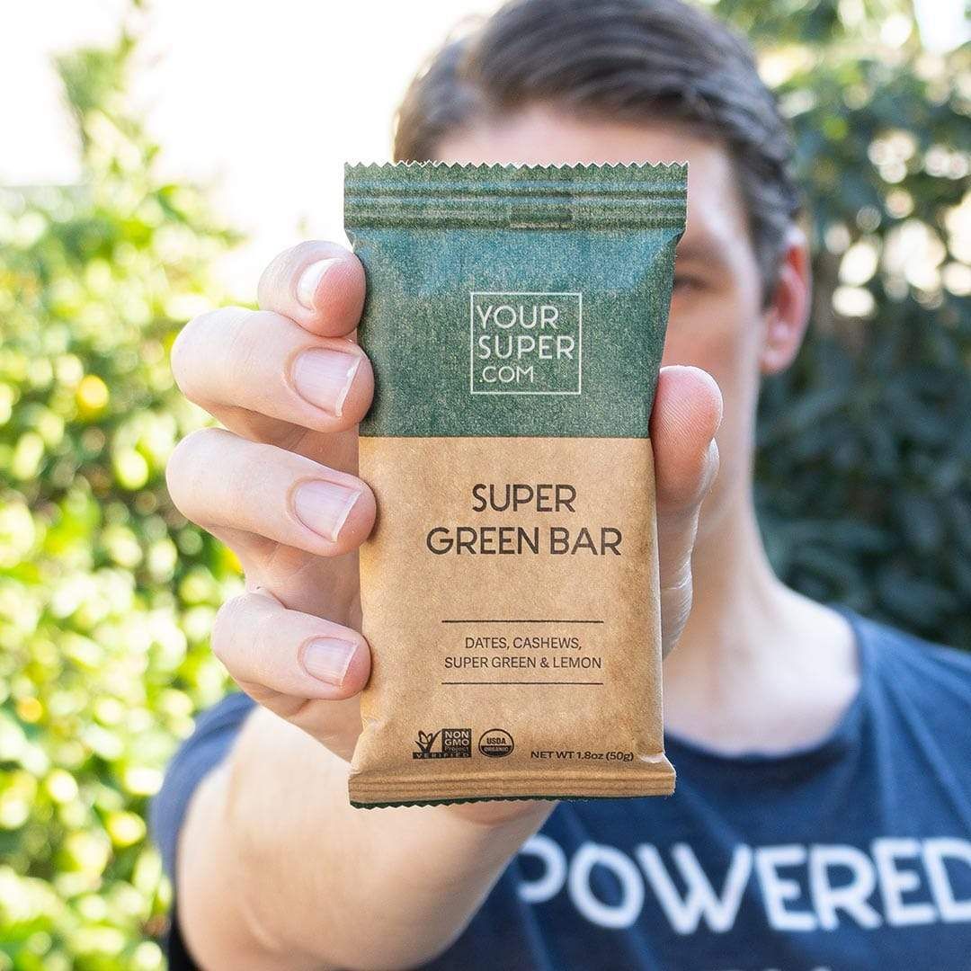 Person hält Super Green Riegel in der Hand. Grün-braune Verpackung mit Text: Super Green Bar, Datteln, Cashews, Super Green & Zitrone. Bio-Siegel.