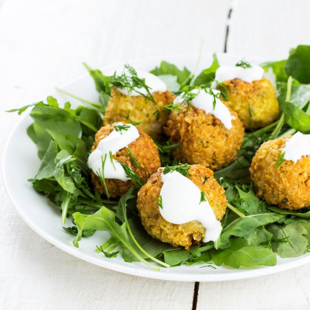 Teller mit sechs Falafelbällchen auf Rucola, mit weißer Sauce und Dill. Helle Holzoberfläche.