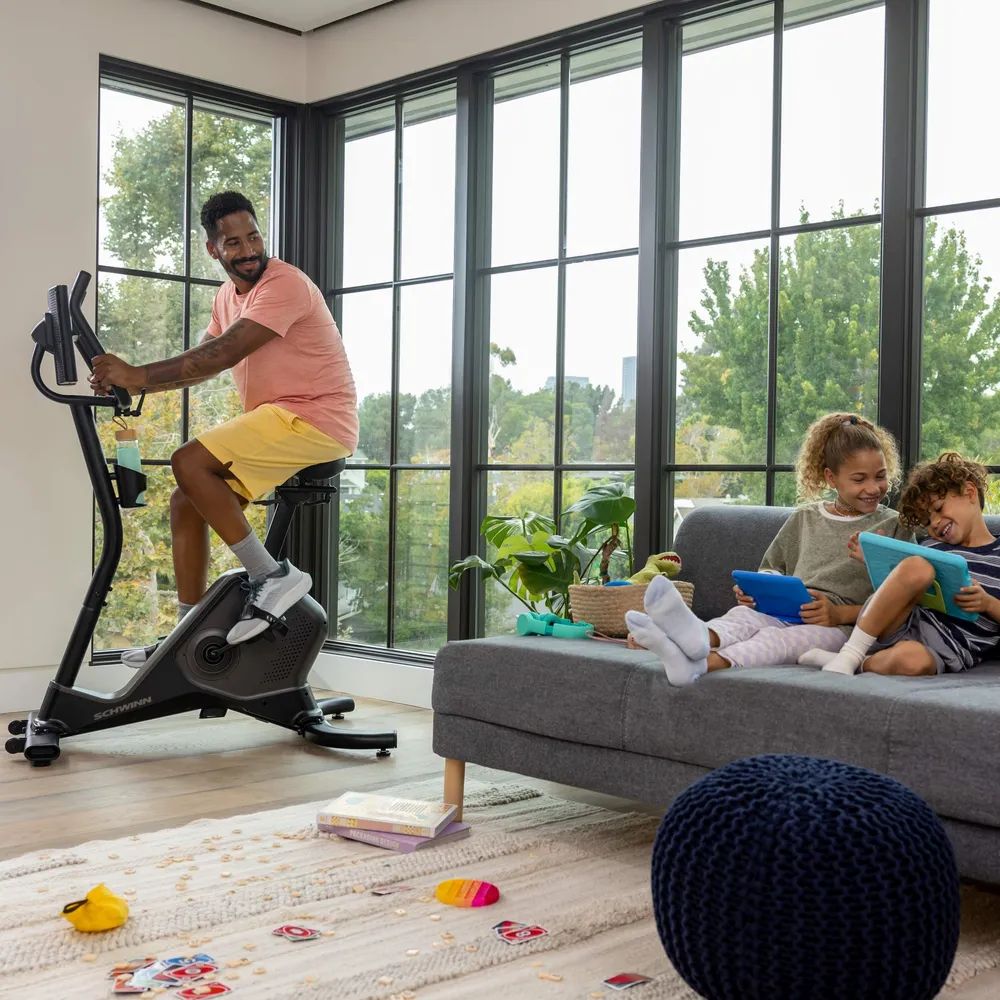 Mann trainiert auf einem Hometrainer. Schwinn-Logo. Kinder auf Sofa. Innenraum mit Fenster.