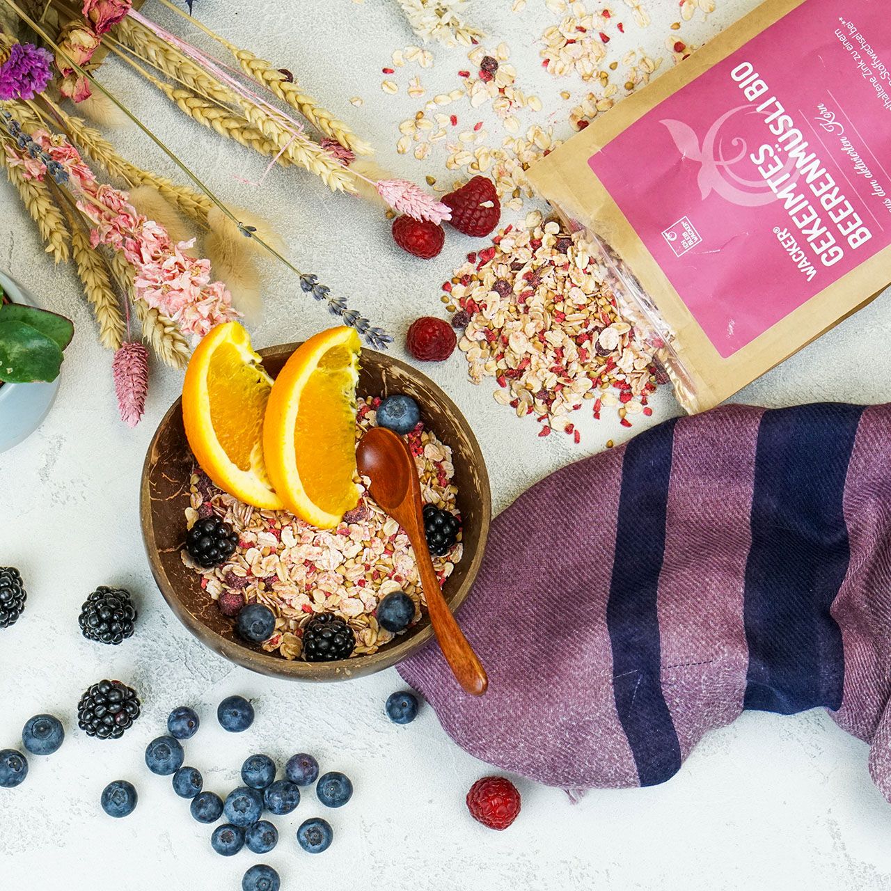 Schale mit Müsli, Blaubeeren, Brombeeren und Orangenscheiben. Im Hintergrund: Verpackung von Wacker Gekeimtes Beerenmüsli Bio.