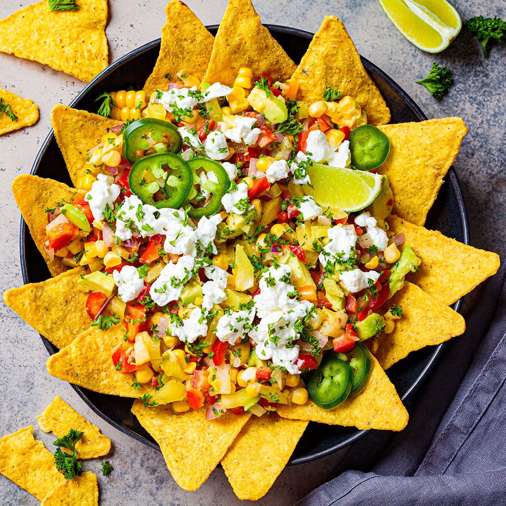 Tortilla-Chips mit Mais, Avocado, Jalapeños und Käse. Auf schwarzem Teller. Limettenspalte.