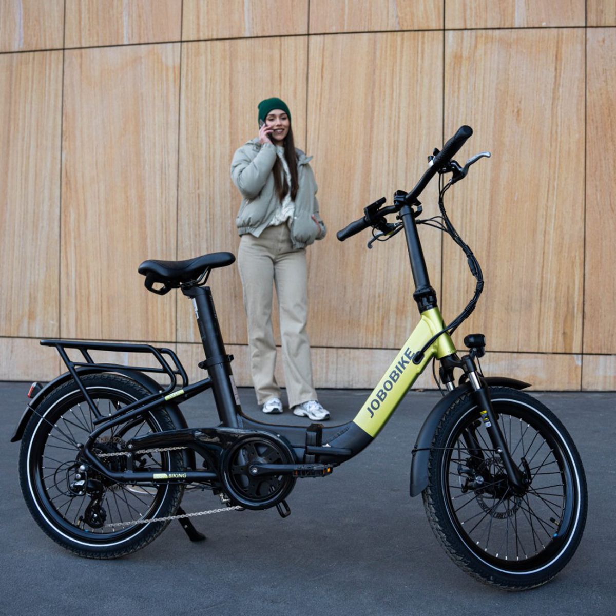 Faltbares E-Bike, grün-schwarz. JOBOBIKE-Logo. Gepäckträger, Schutzbleche. Person telefoniert neben dem Fahrrad.