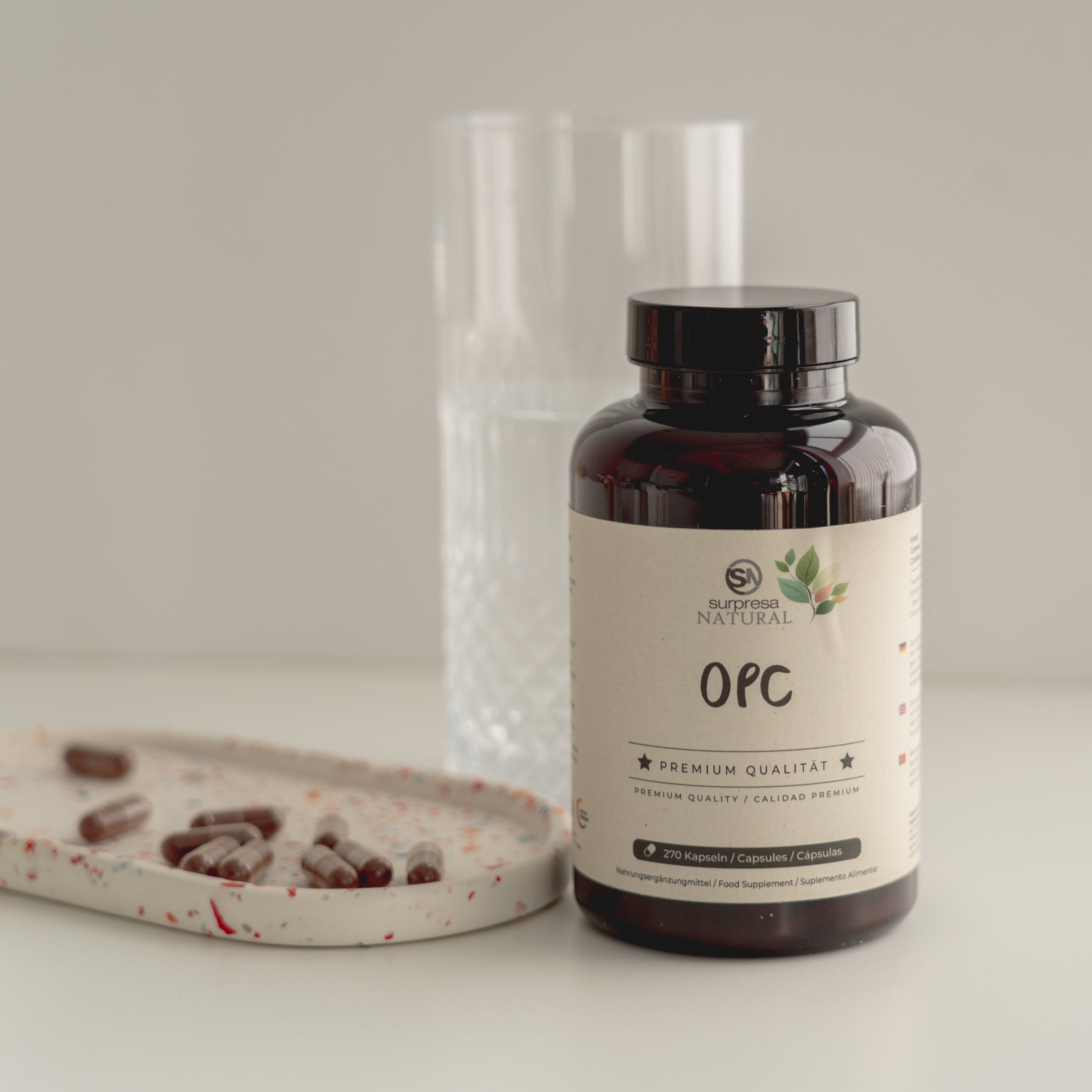 Flasche mit OPC Kapseln, Kapseln auf einem kleinen Teller, ein Glas im Hintergrund.
