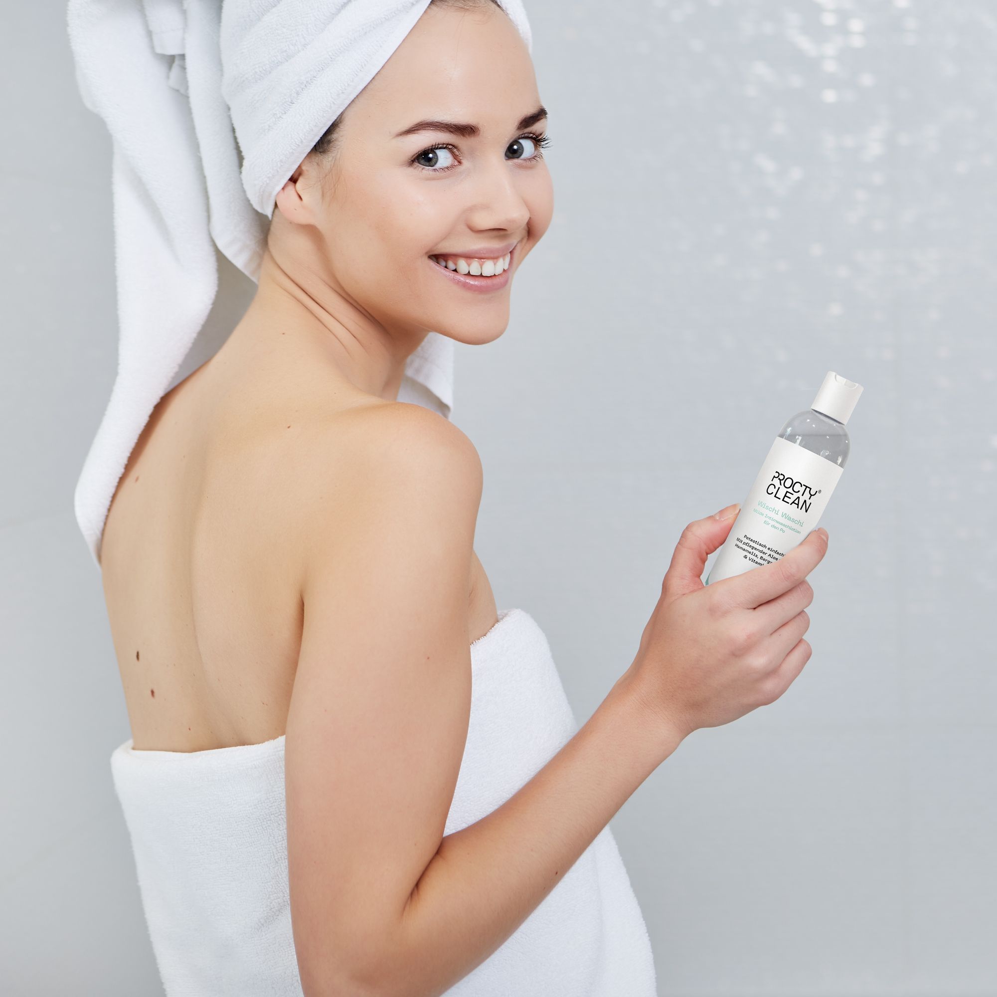Frau mit Handtuch hält Produktflasche. Hintergrund: Badezimmer.