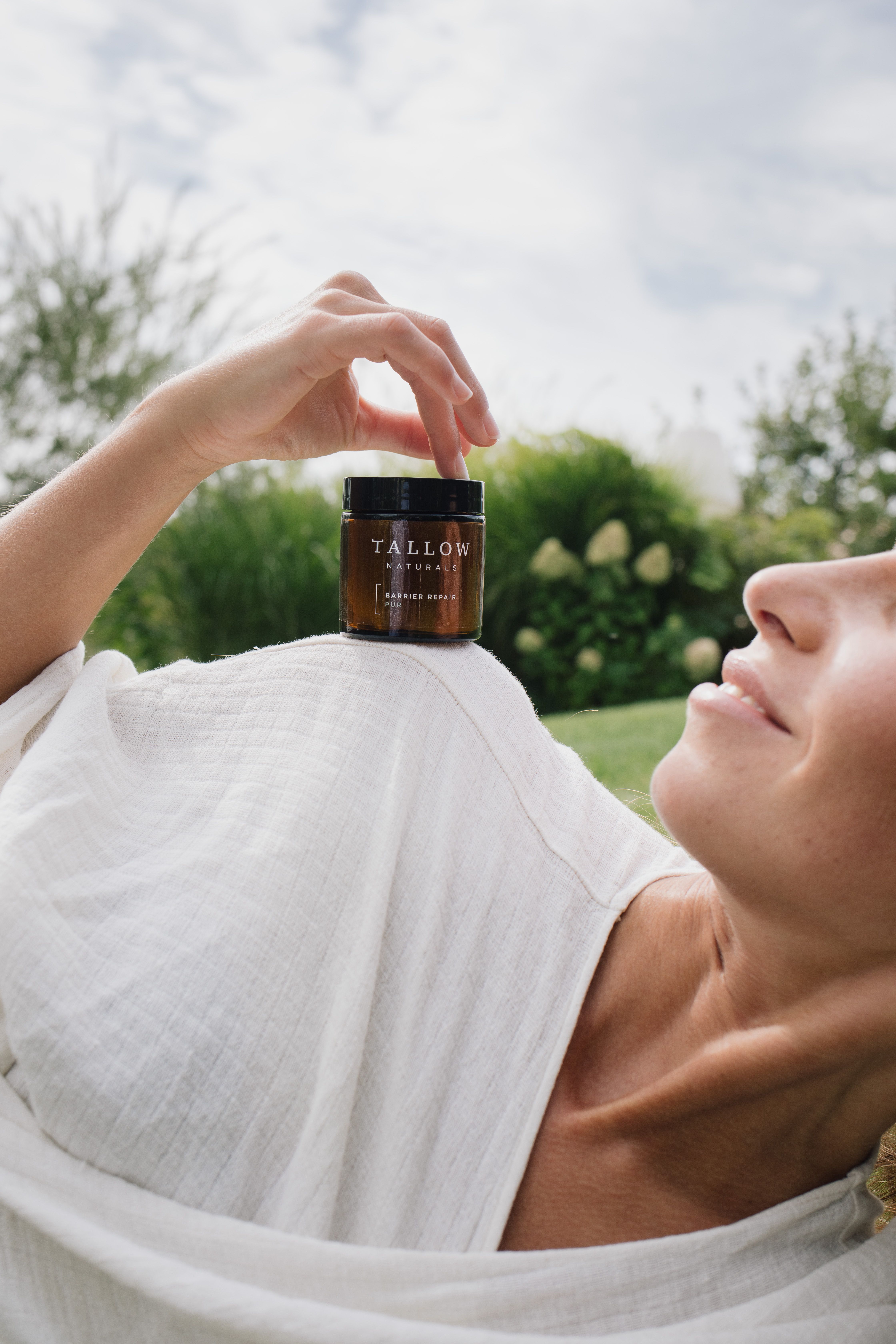 Frau liegt im Freien und hält eine braune Dose mit schwarzem Deckel. Aufschrift: TALLOW NATURALS. Produktname nicht lesbar.