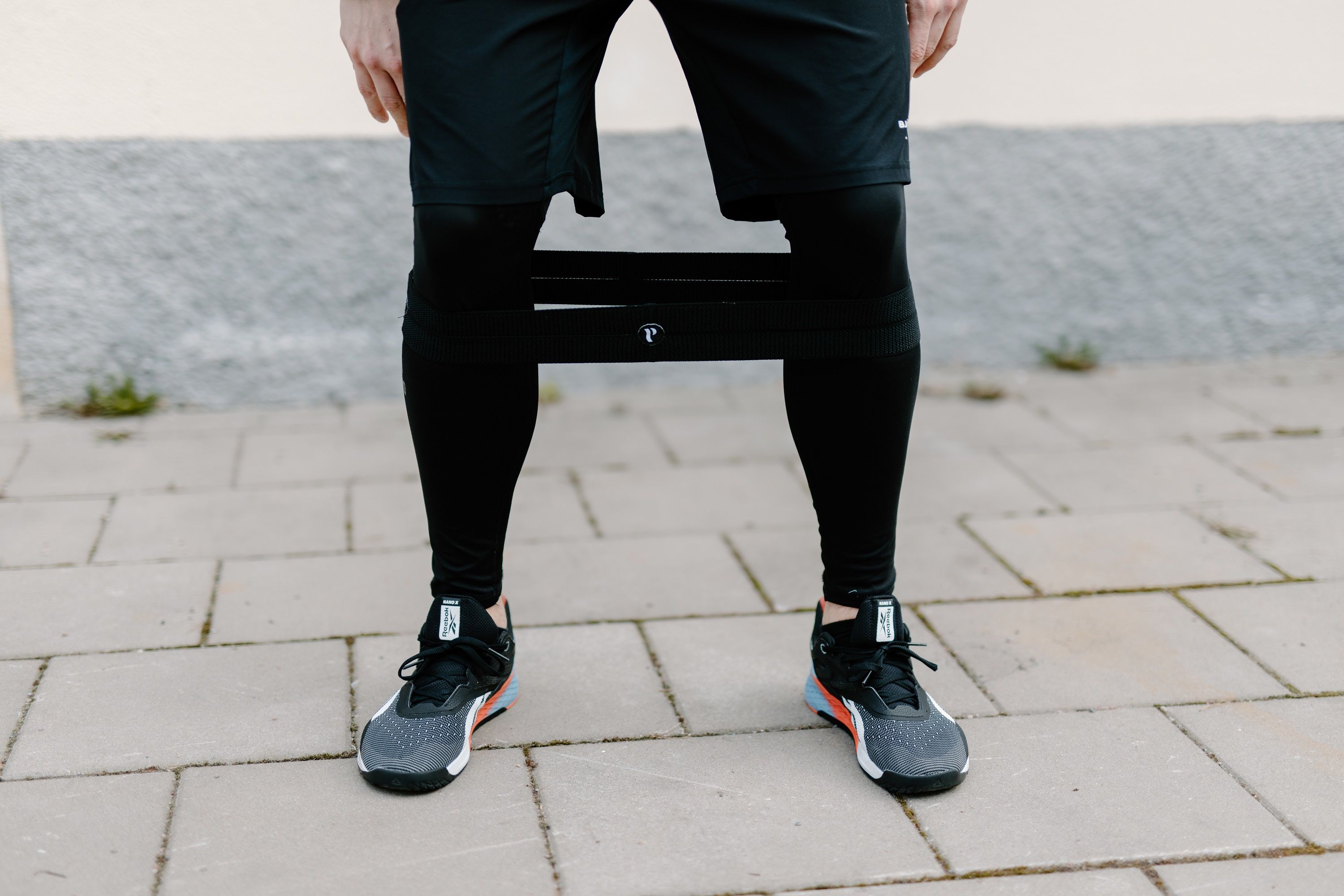 Person mit schwarzer Beinbandage. Schwarze Hose, Turnschuhe. Pflastersteine im Hintergrund.