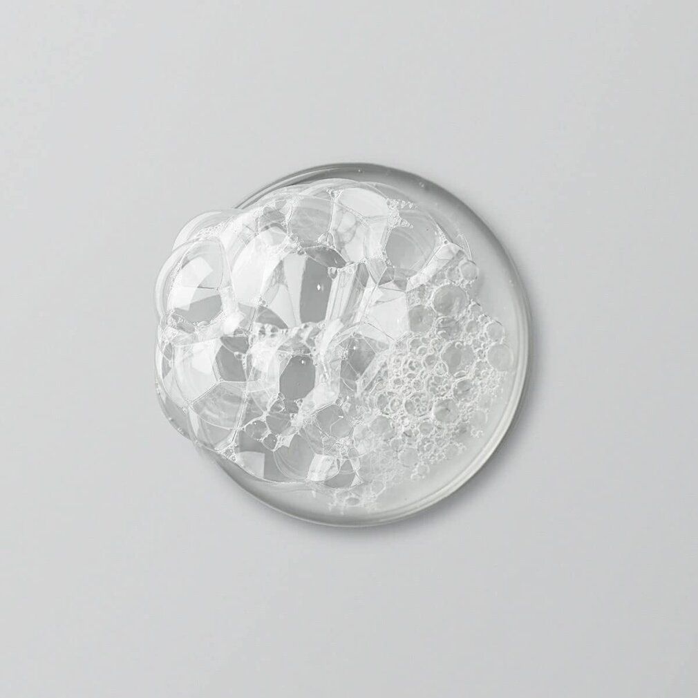 Klare Flüssigkeit mit Schaumblasen in einer runden, durchsichtigen Schale. Draufsicht auf hellem Hintergrund.