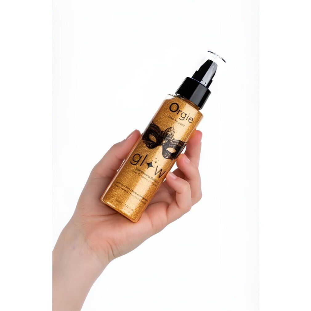 Goldfarbene Körperöl-Flasche in Hand gehalten. Schwarzer Pumpverschluss. Produktname und Masken-Design. Text: Glow, Shimmering Body Oil.
