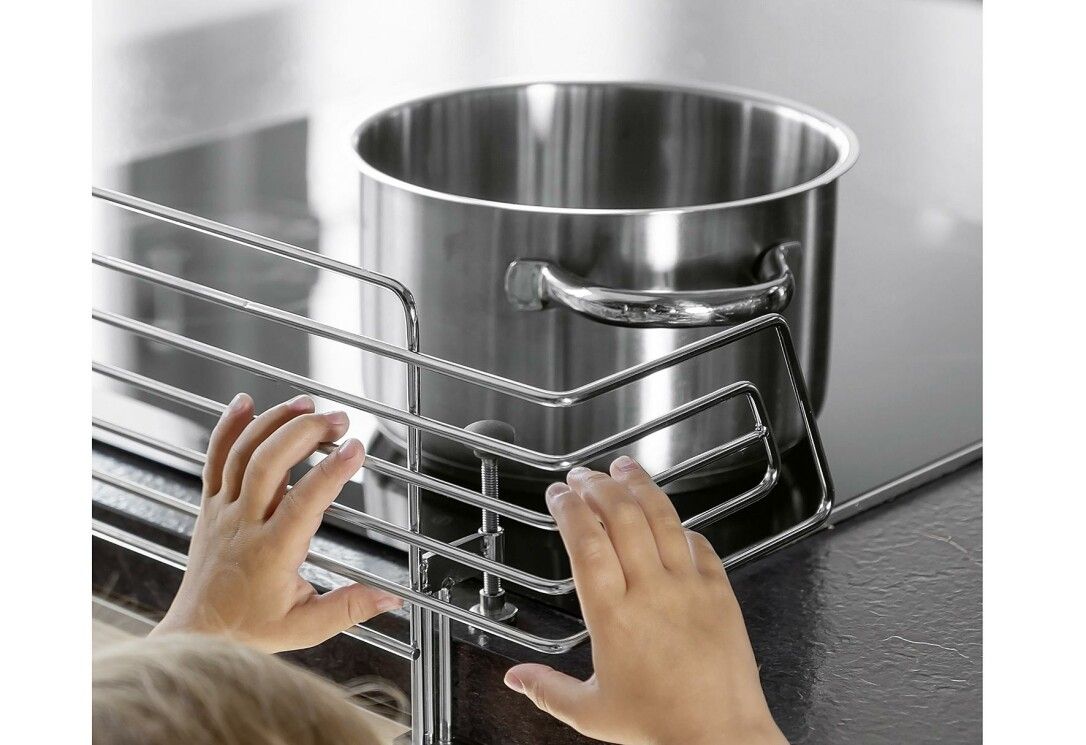 Metallgitter vor einem Kochfeld mit einem Topf. Hände eines Kindes berühren das Gitter.