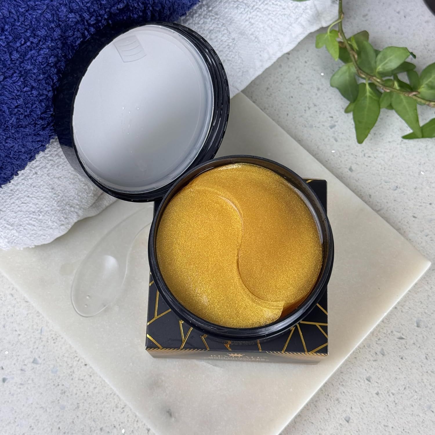 Offener Tiegel mit goldenen Augenpads und Applikator. Auf weißer Oberfläche.