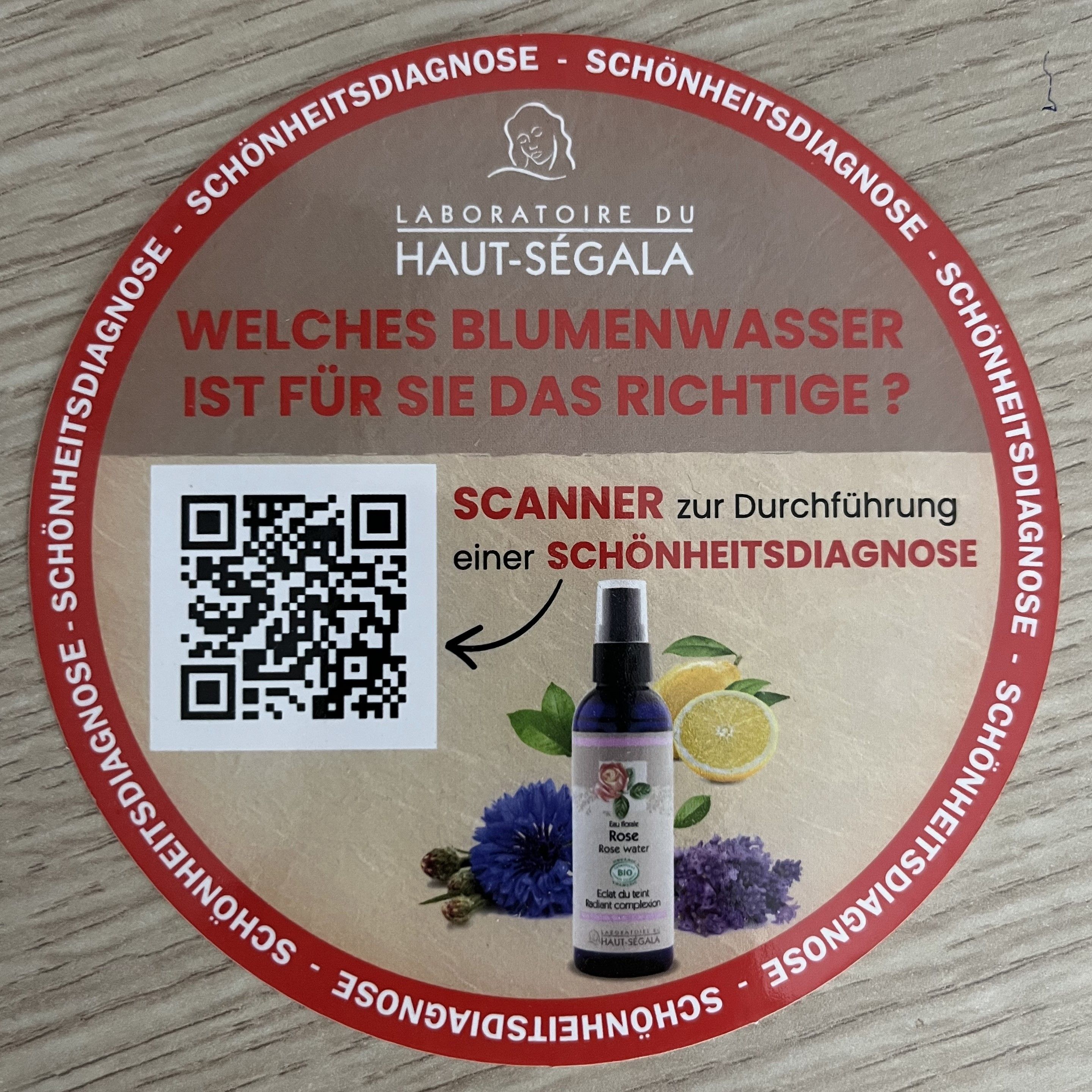 Rundes Werbeschild mit Produkt und QR-Code. Text: Laboratoire du Haut-Ségala, Geraniumwasser. Mit Blüten und Zitrone.
