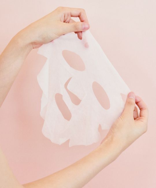 Weiße Gesichtsmaske mit Aussparungen für Augen, Nase und Mund. Gehalten von zwei Händen vor rosa Hintergrund.
