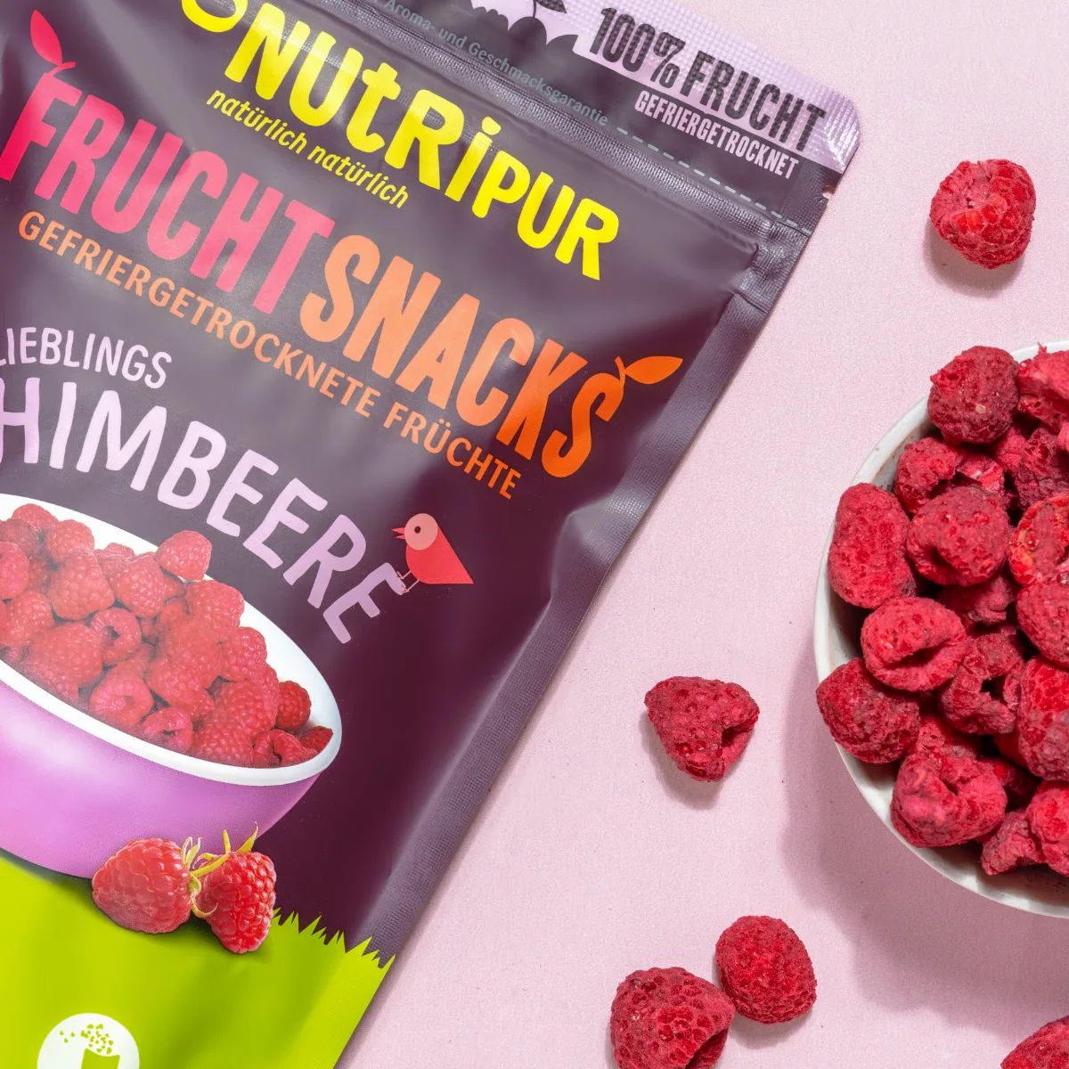 Nahaufnahme von NutriPur Fruchtsnacks Himbeeren Beutel. Daneben Schüssel mit Himbeeren. Einzelne Himbeeren.