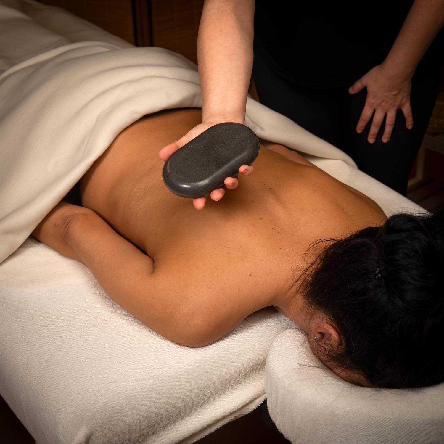Großer, ovaler Stein wird auf dem Rücken gehalten. Person liegt auf einer Liege, bedeckt mit einem weißen Tuch. Helle Haut.