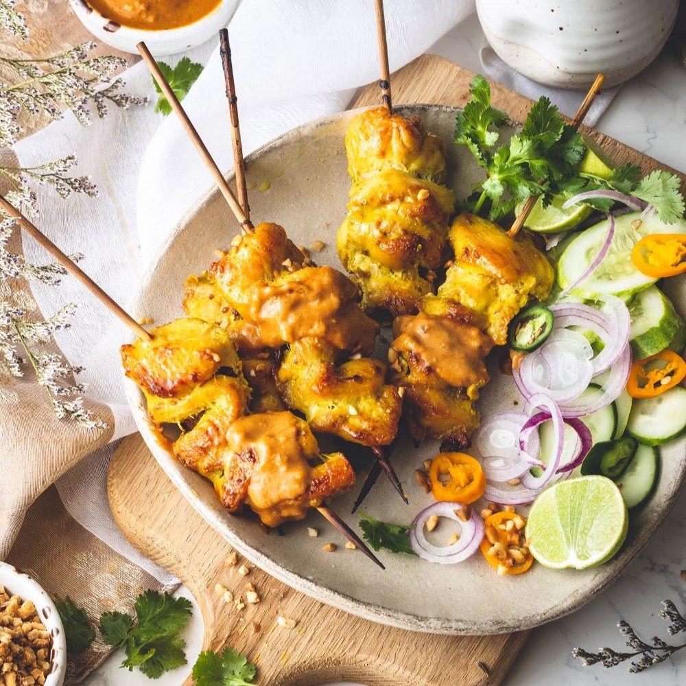 Satay-Spieße auf einem Teller mit Beilagen. Helle Soße in einer Schale. Holzbrett und Dekoration.
