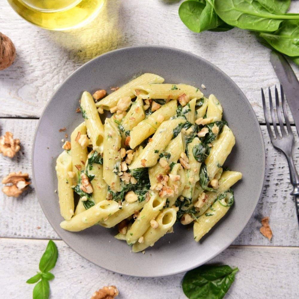Pasta mit Spinat und Walnüssen auf grauem Teller. Draufsicht, umgeben von Zutaten und Besteck.