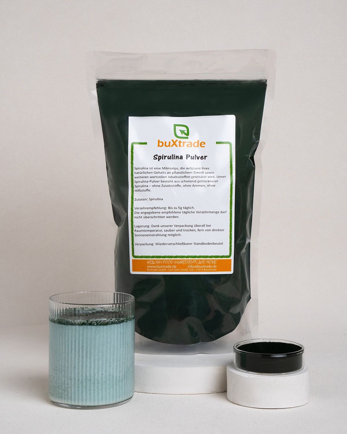 Beutel mit Spirulina Pulver, Glas mit Flüssigkeit, kleiner Behälter. Aufdruck: buXtrade, Spirulina Pulver.