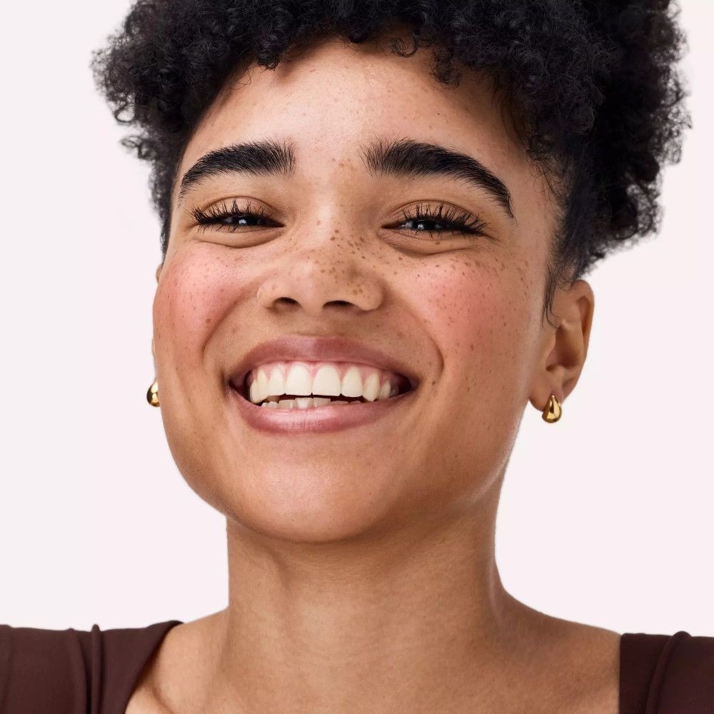 Nahaufnahme einer lächelnden Frau mit Rouge auf den Wangen. Sie trägt goldene Ohrringe.