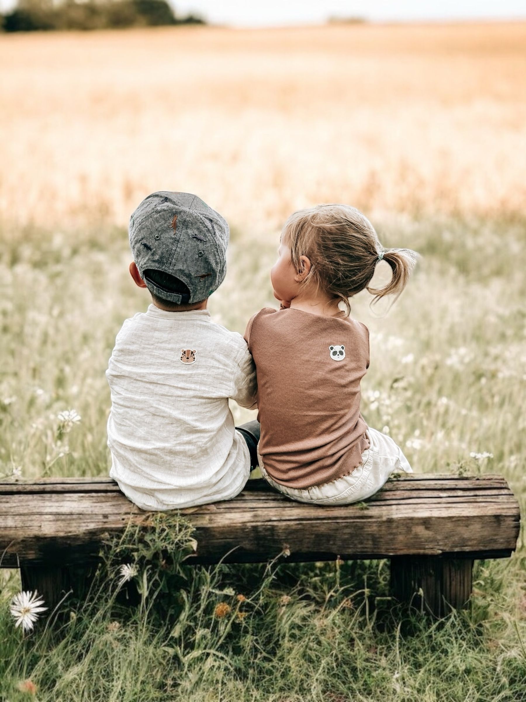 Zwei Kinder sitzen auf einer Holzbank, Rücken zur Kamera. Auf ihren Shirts sind Tiergesichter aufgedruckt. Grüne Wiese im Hintergrund.