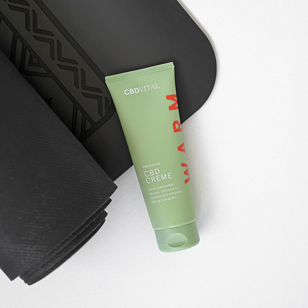 Grüne Tube CBD VITAL Wärmende CBD Creme neben einer aufgerollten Yogamatte.